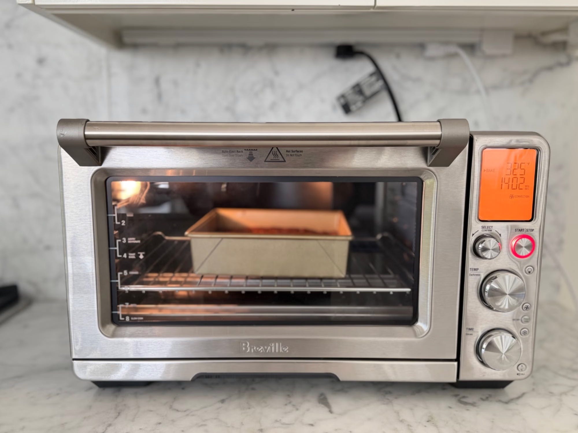 Breville countertop toaster oven baking a loaf pan on a marble kitchen counter.