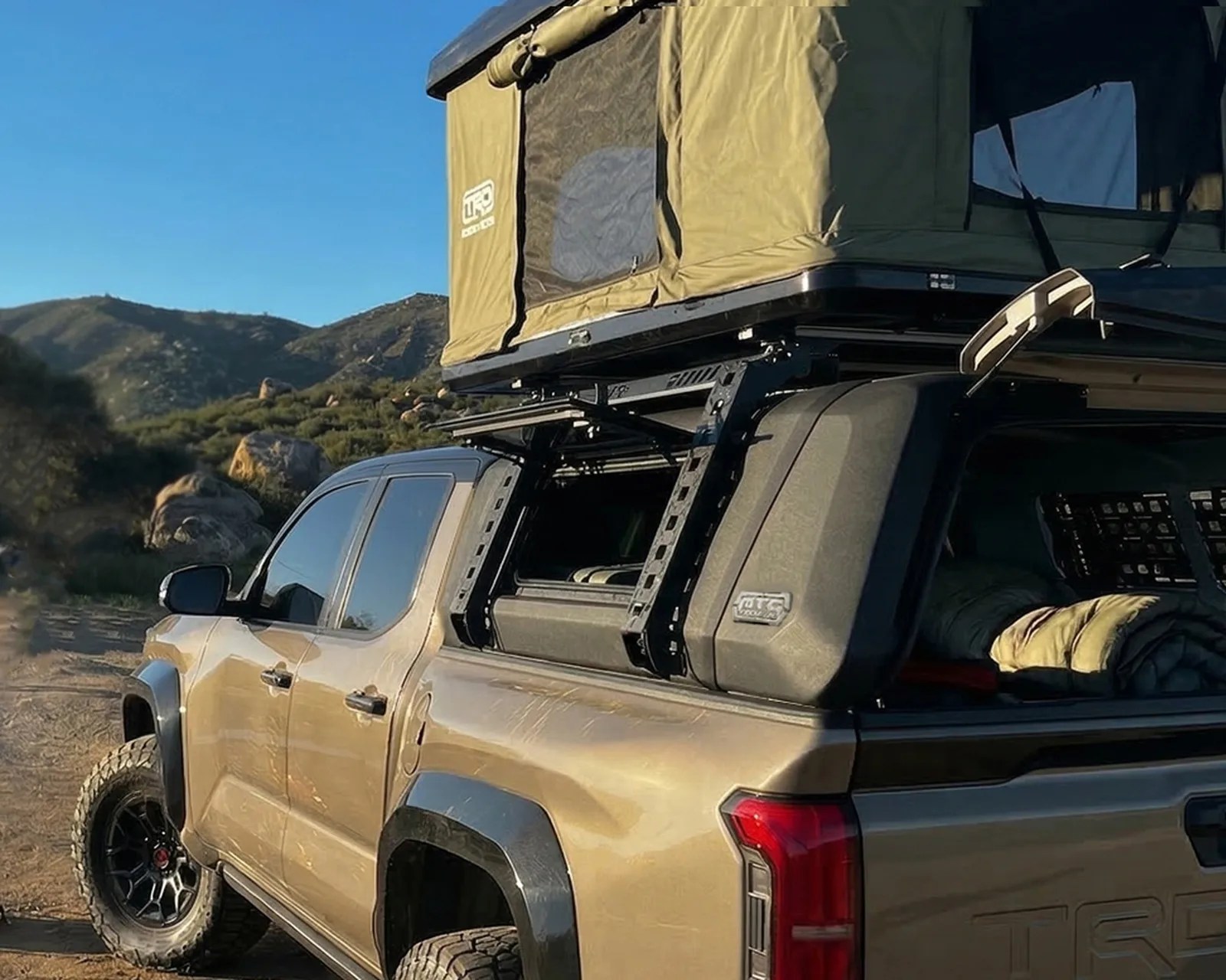 Tan pickup truck with off-road tires and a rooftop tent mounted on a black rack in a mountainous outdoor setting.