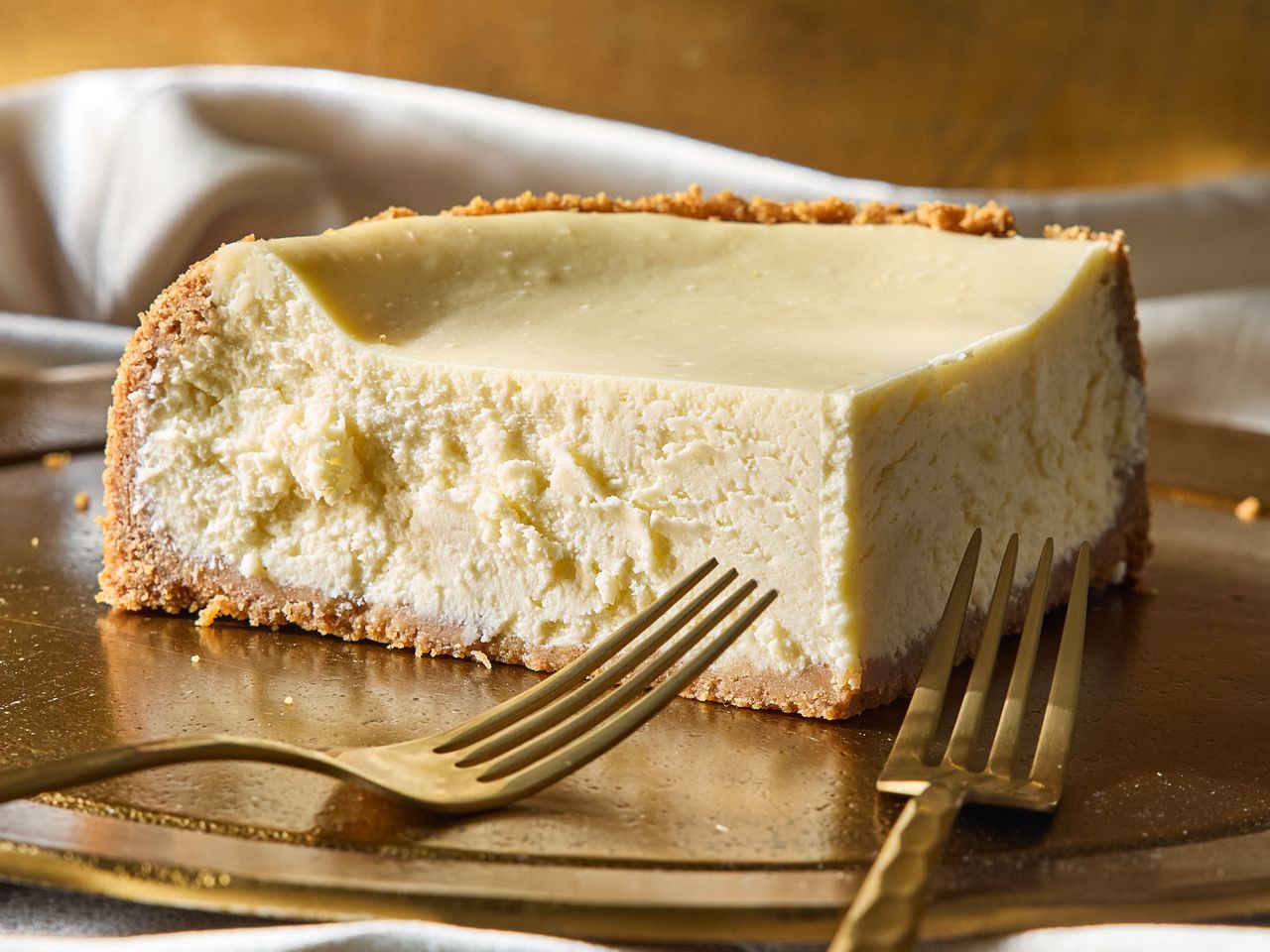 A slice of cheesecake on a gold surface with two forks nearby