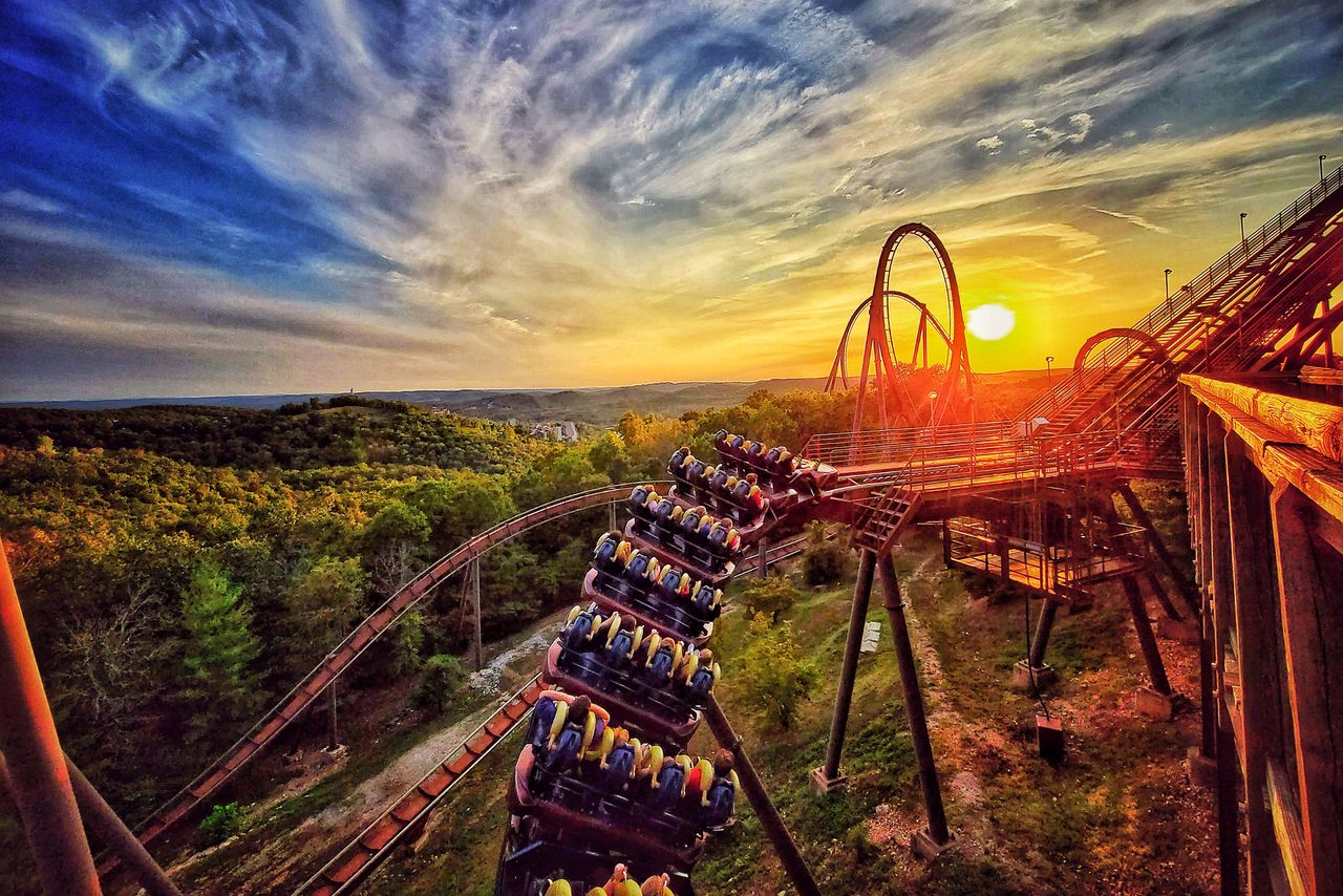 A roller coaster at sunset riders descending a slope against a scenic wooded landscape with a looping track in view