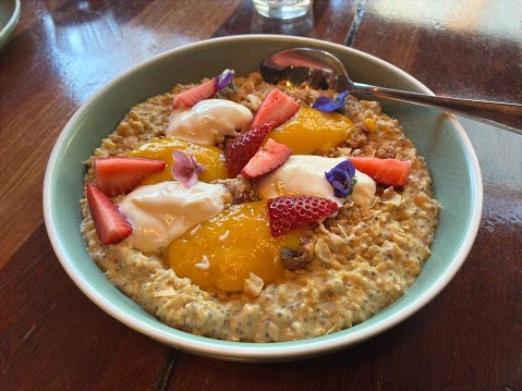 A bowl of chia oatmeal topped with yogurt, peach slices, strawberries, and edible flowers on a wooden table