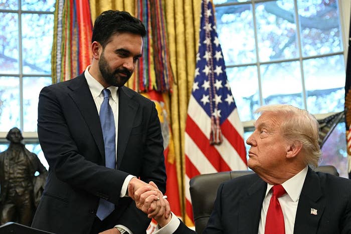 Two men in formal attire shaking hands in an office with flags and a statue visible in the background