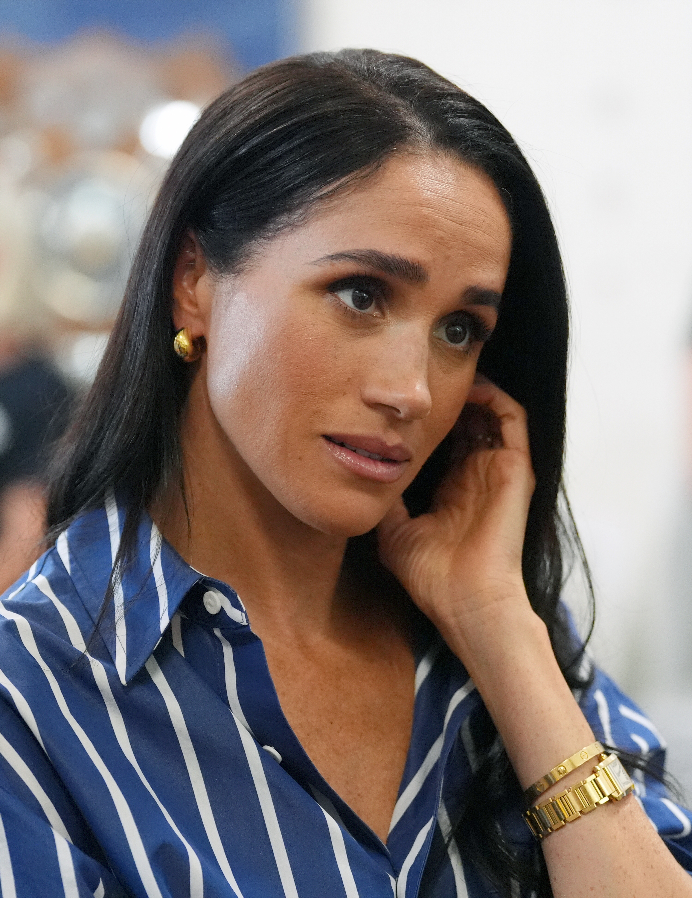 Meghan, Duchess of Sussex, listens intently while touching her ear, wearing a blue and white striped shirt and gold jewelry.