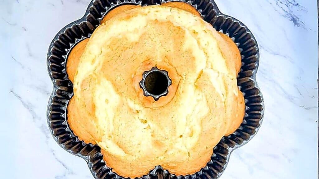 A golden-brown bundt cake rests in a fluted metal baking pan on a white marble surface, viewed from above.
