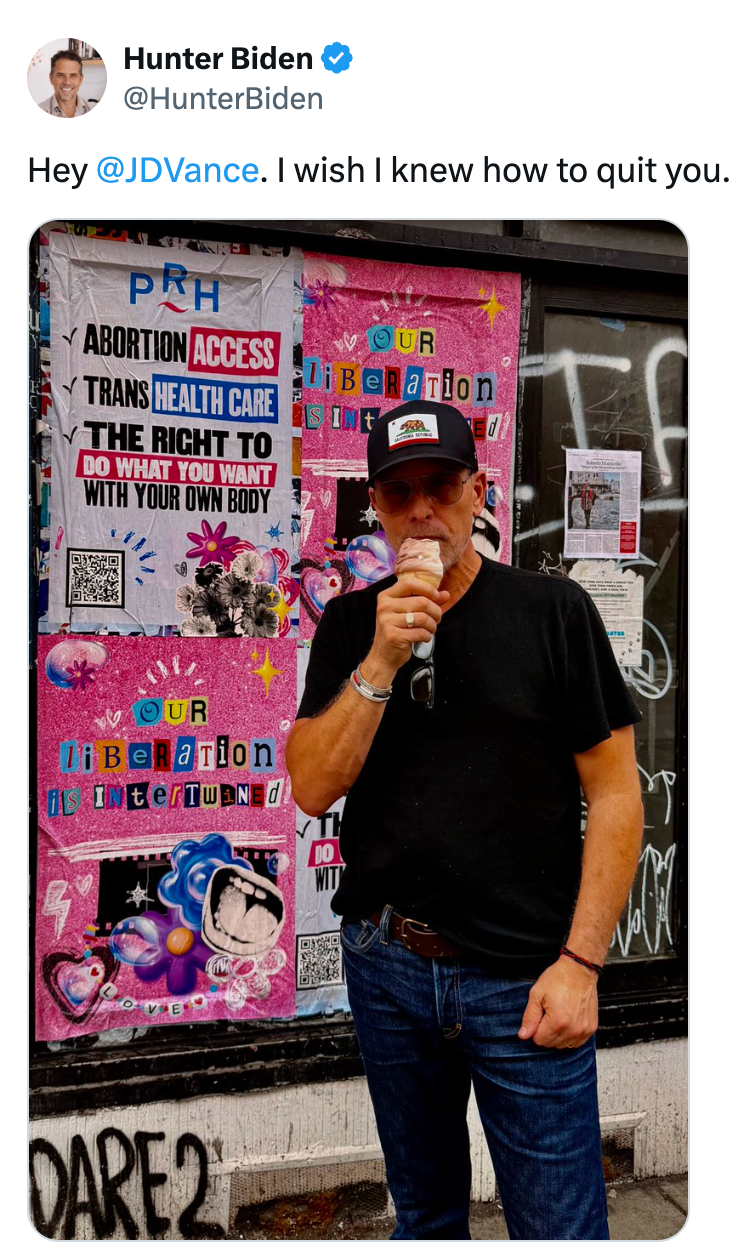 Hunter Biden stands in front of a street art poster, casually dressed, enjoying an ice cream cone