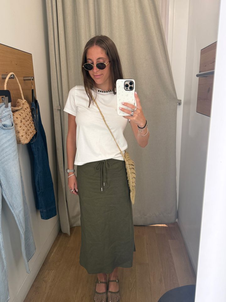 Person in a dressing room trying on an outfit wearing a white top and green skirt holding a phone for a mirror selfie