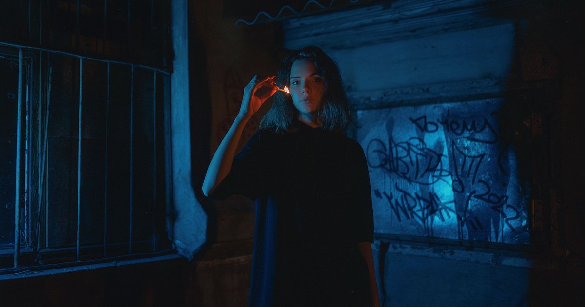 A woman stands in a dimly lit alley holding a burning stick, surrounded by graffiti.