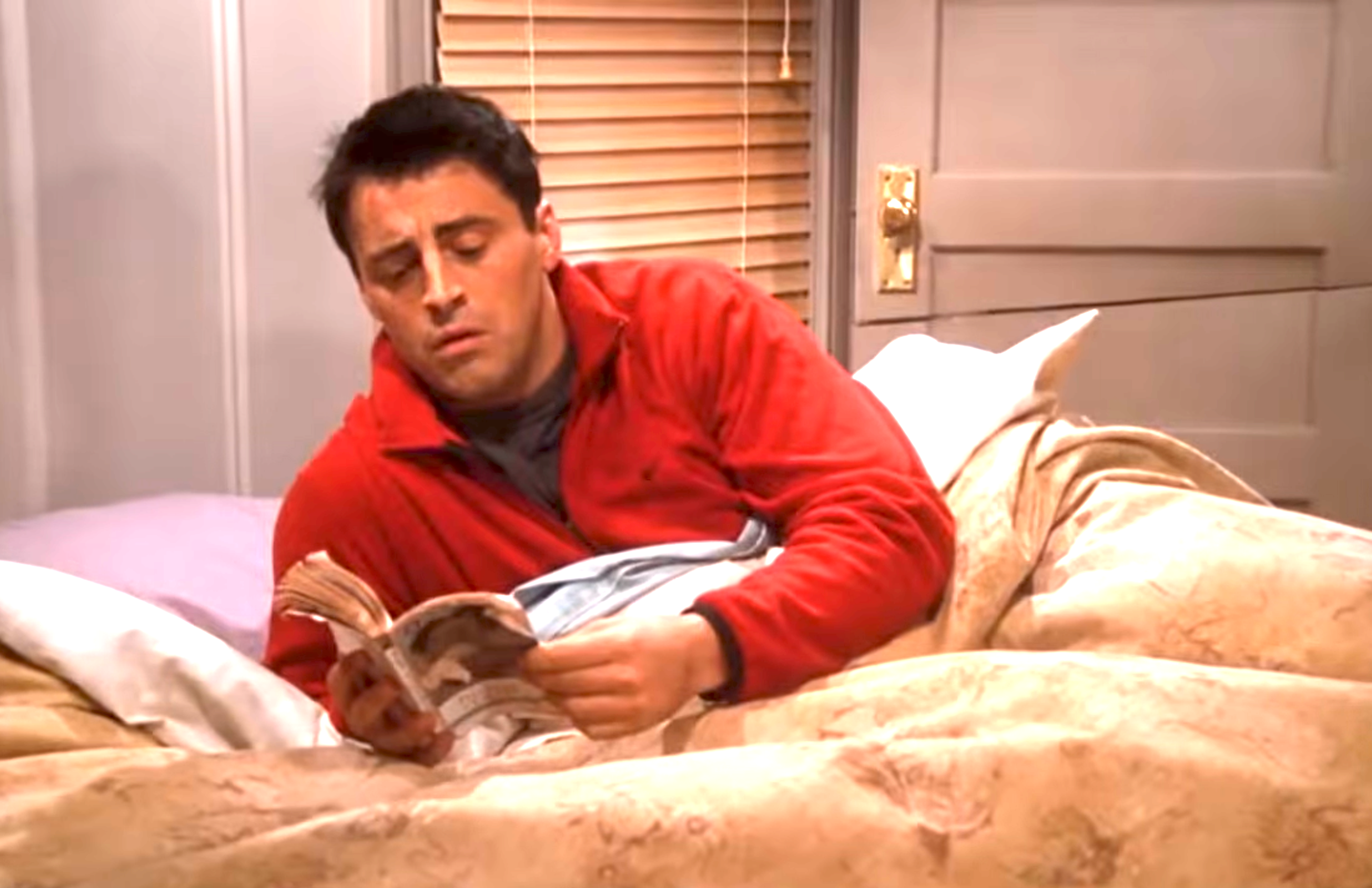 Person in a cozy bed reading a magazine, wearing a casual red jacket, looking engaged