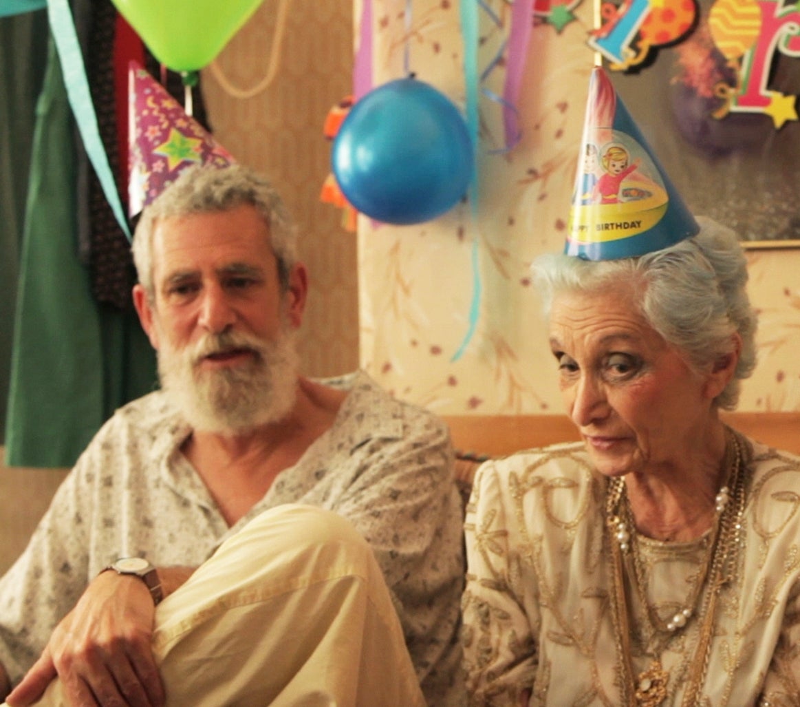 Three people in party hats sit together at a celebration. The older woman looks thoughtfully forward, while the man on her right gestures animatedly
