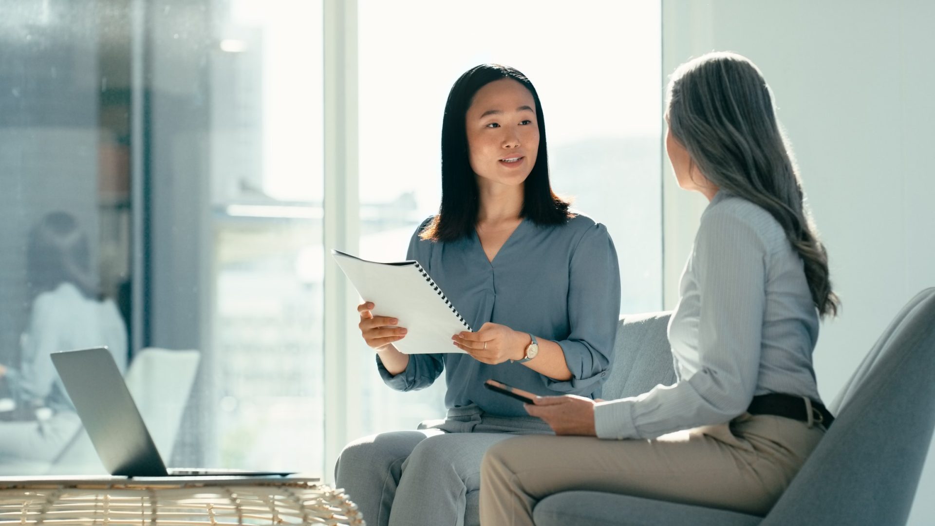 Discover two businesswomen sit together in an office or conference room discussing papers next to laptop