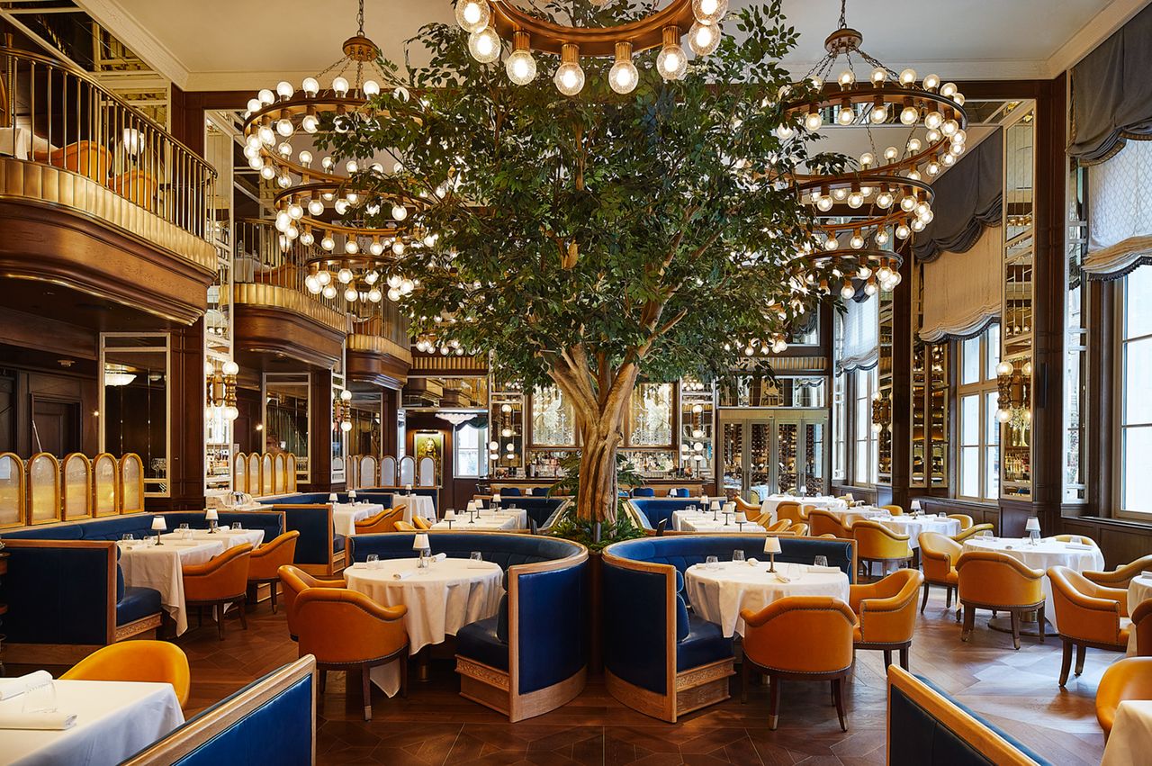 Elegant restaurant interior with round tables blue booths and a central tree under chandeliers
