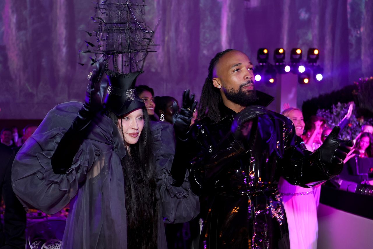 Two individuals posing at an event one wearing a creative hat and outfit and the other dressed in shiny clothing with people visible in the background