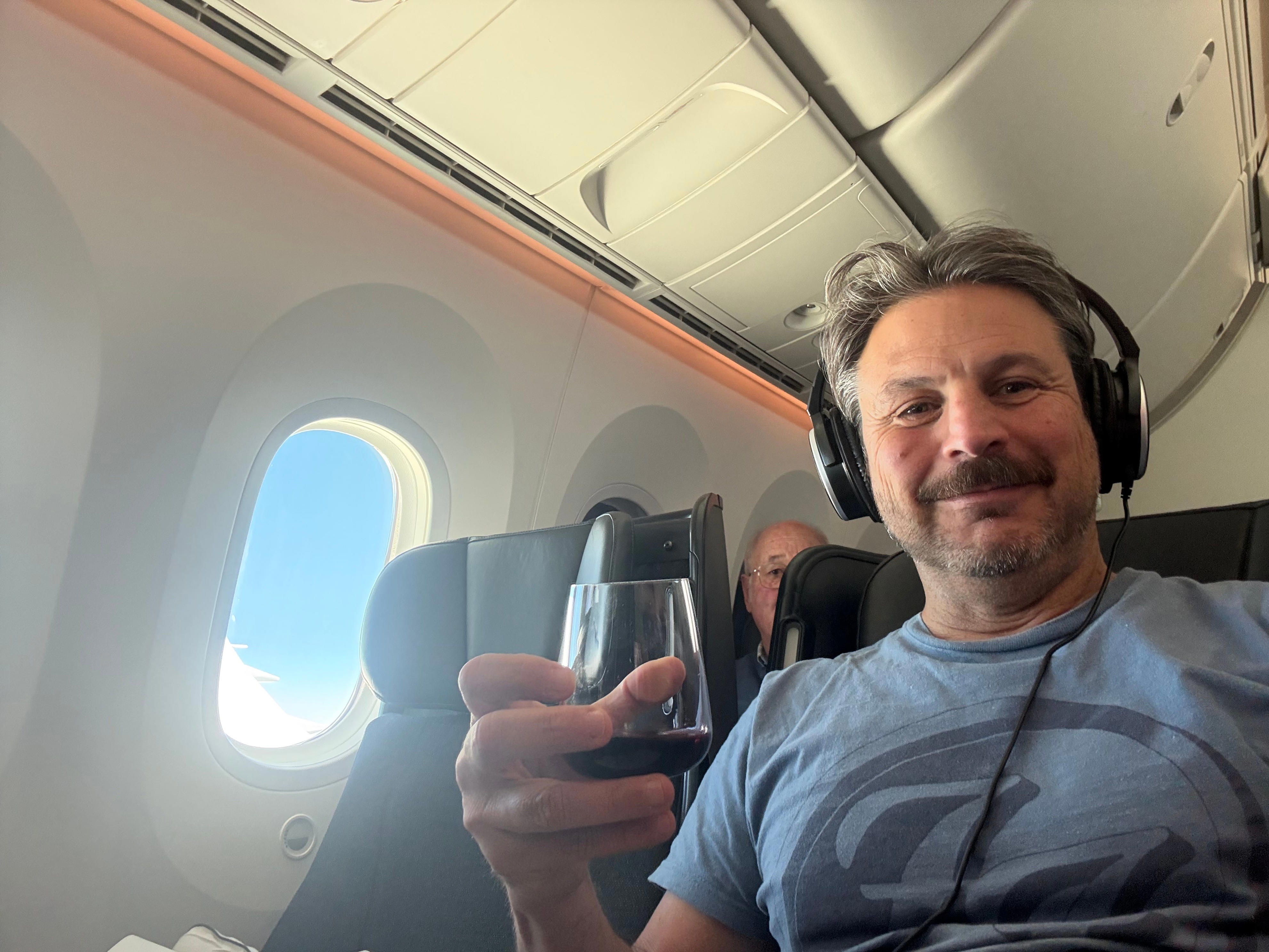 Author Ash Jurberg in premium economy seat with drink, smiling , wearing headphones