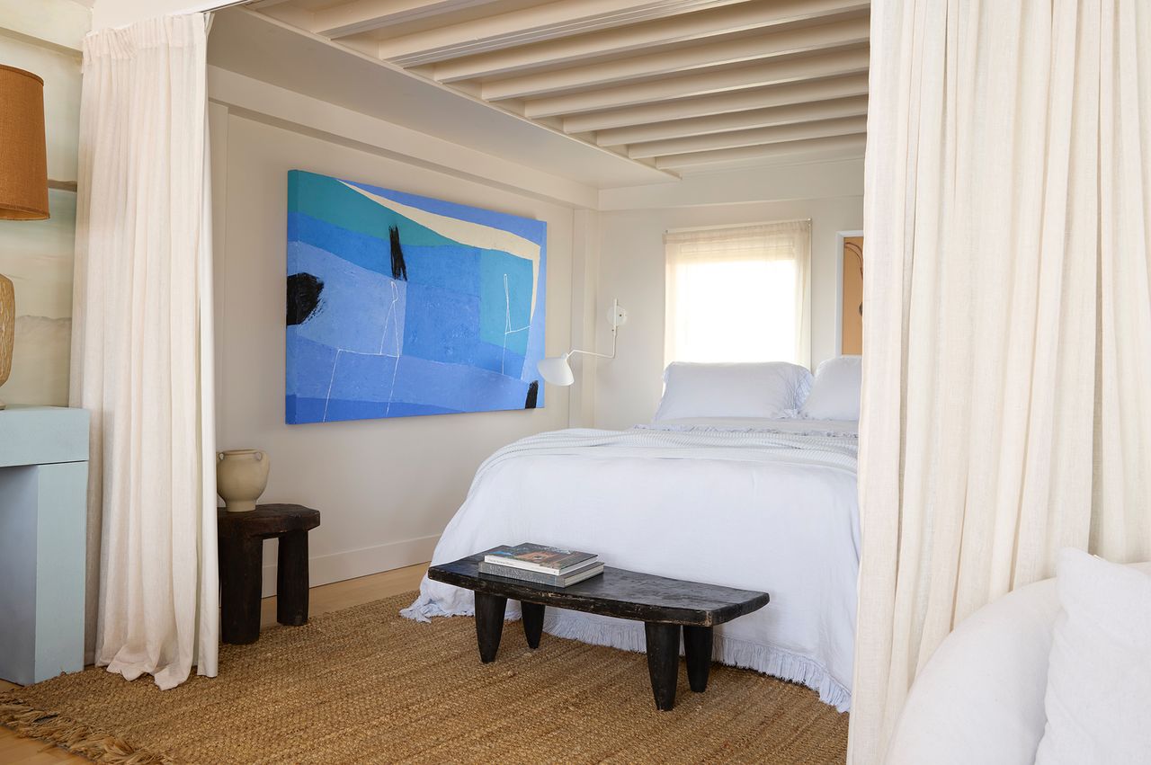 A decorated guest room with a bed a blue abstract painting on the wall and natural decor elements like a woven rug and curtains