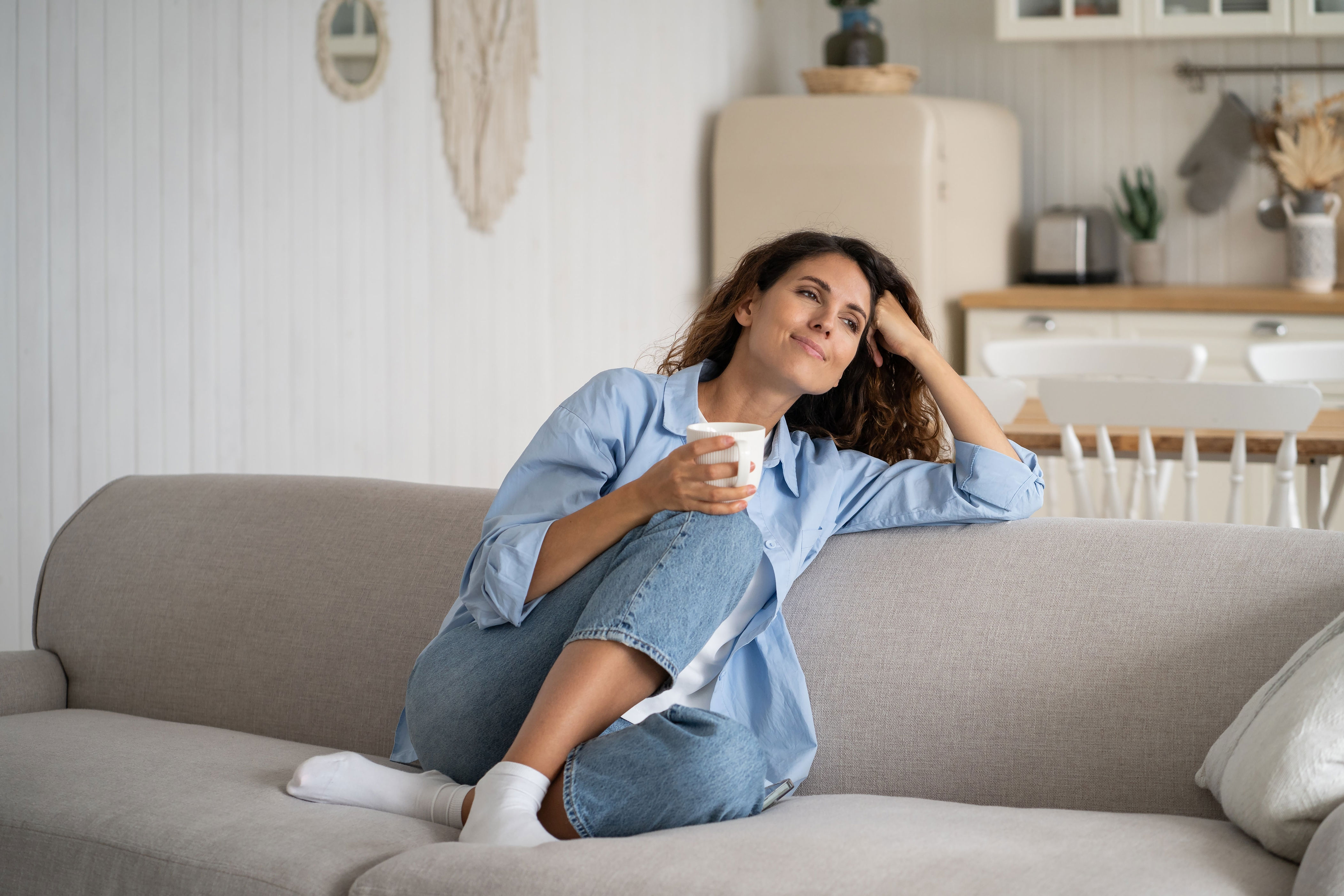 Person relaxing on a couch with a drink, looking thoughtfully into the distance, wearing casual jeans and a shirt. Cozy home setting