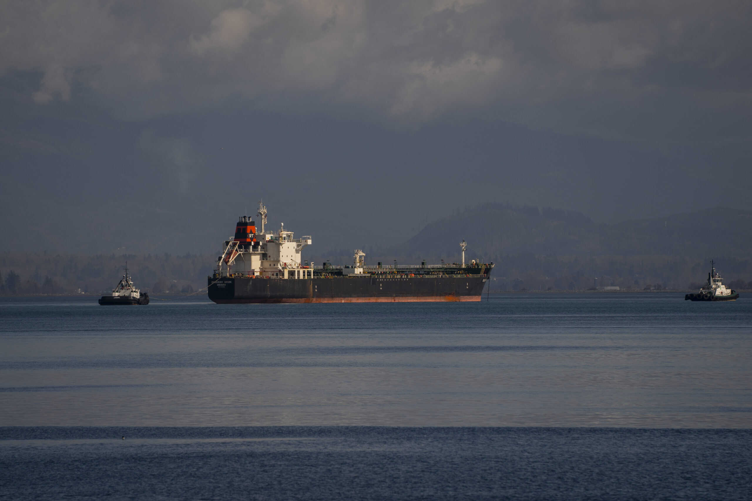 A large, dark-hulled oil tanker with a prominent white superstructure and orange-red funnel, bearing an 'RT' logo, is centrally positioned in the frame, navigating calm dark blue-green waters. Two white and dark grey tugboats flank the tanker, one on the left pulling a line and one on the right, assisting its movement. Hazy, forested mountains extend across the distant horizon under a dramatic, overcast sky with subtle light variations.