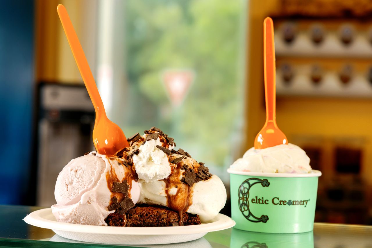 Two servings of ice cream one on a plate with chocolate toppings and one in a Celtic Creamerybranded cup with spoons