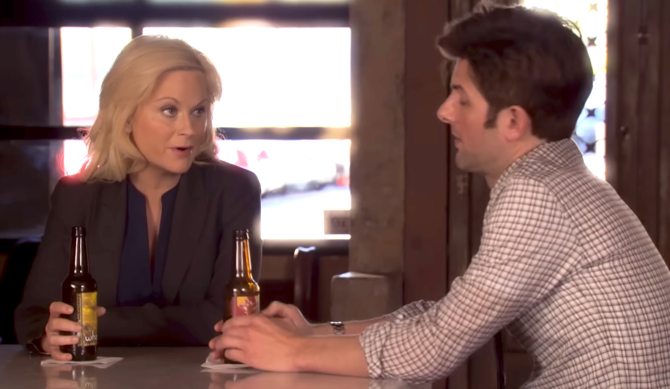 Two people sitting at a bar, engaged in conversation, each holding a beer bottle