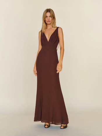Person in a sleeveless, floor-length gown with a deep V-neck, standing in a neutral setting, showcasing elegant, minimalist fashion style