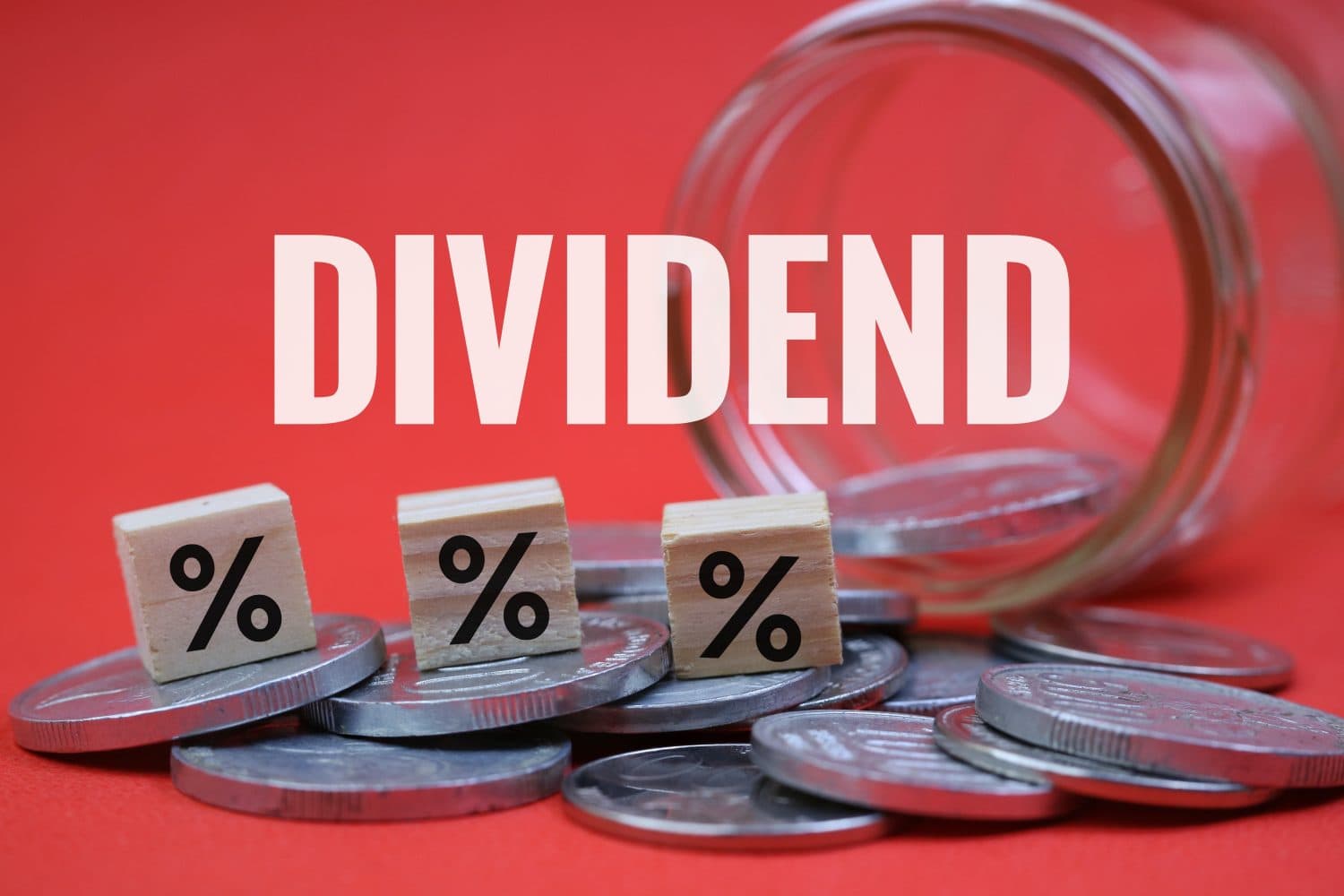The word 'DIVIDEND' in large white letters against a red background. Below it, three small wooden blocks with black percentage symbols rest on stacks of silver coins. A clear glass jar is partially visible in the upper right, tipped over with more silver coins around it.
