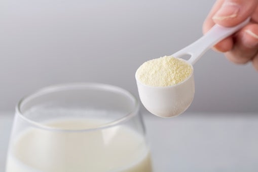 A hand holds a scoop of powdered milk above a glass of liquid, suggesting preparation or addition of the powder to the drink