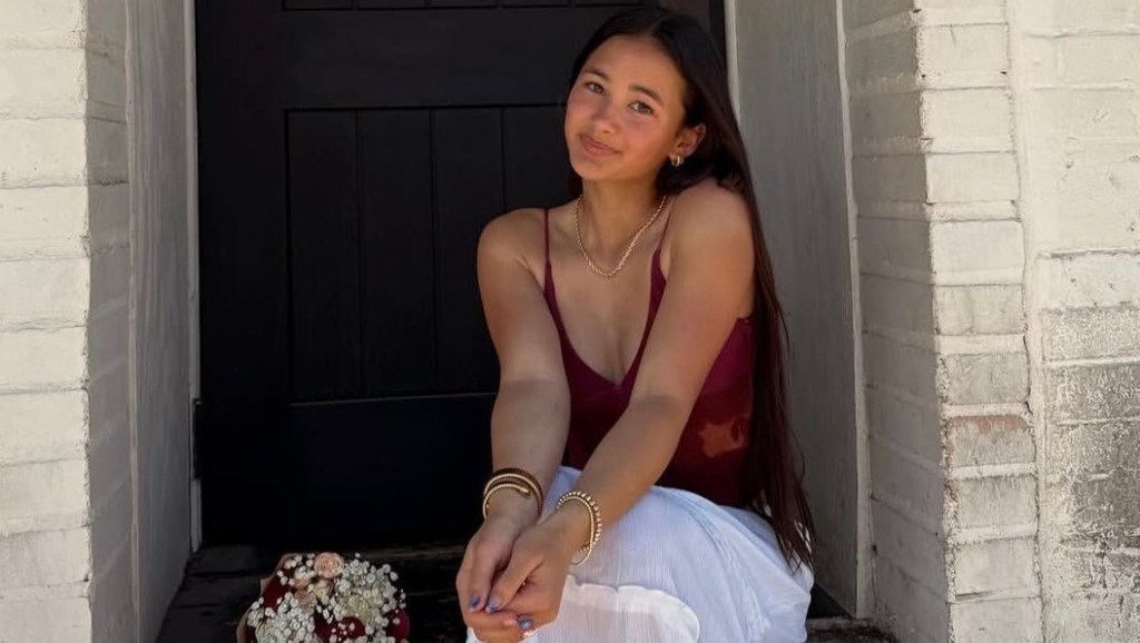 Kaylee Hottle, a young woman with long dark hair, wearing a red top and white skirt, seated on a step with a bouquet of flowers next to her.