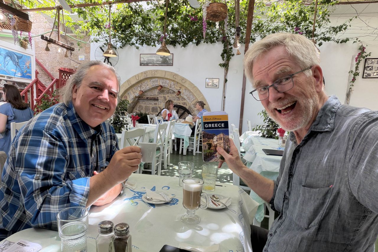 Rick Steves seated at a table in a restaurant holding a guidebook another man seated across with food and drinks