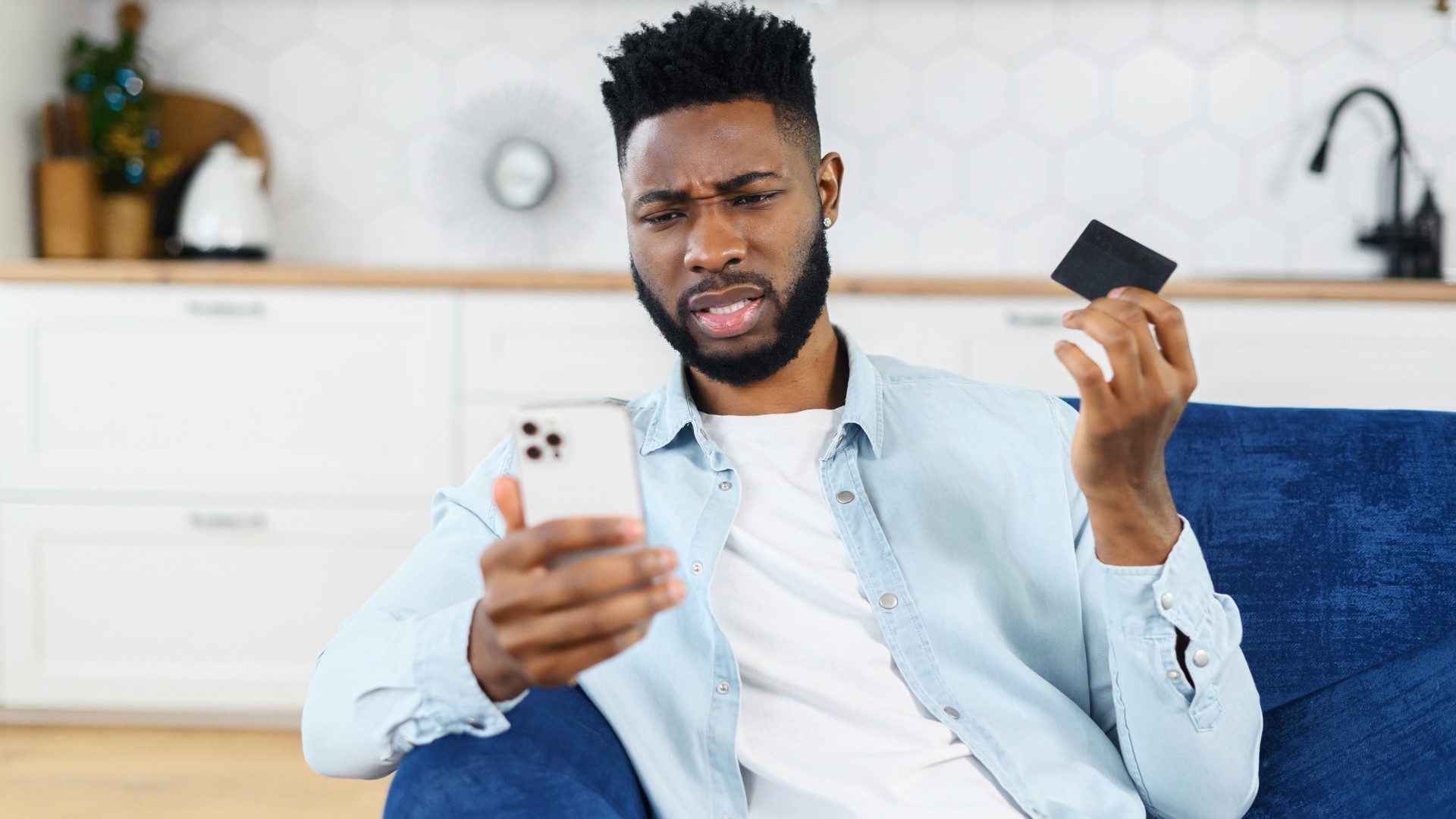 Discover an irritated young man holding his smartphone and a credit or debit card sits on a couch