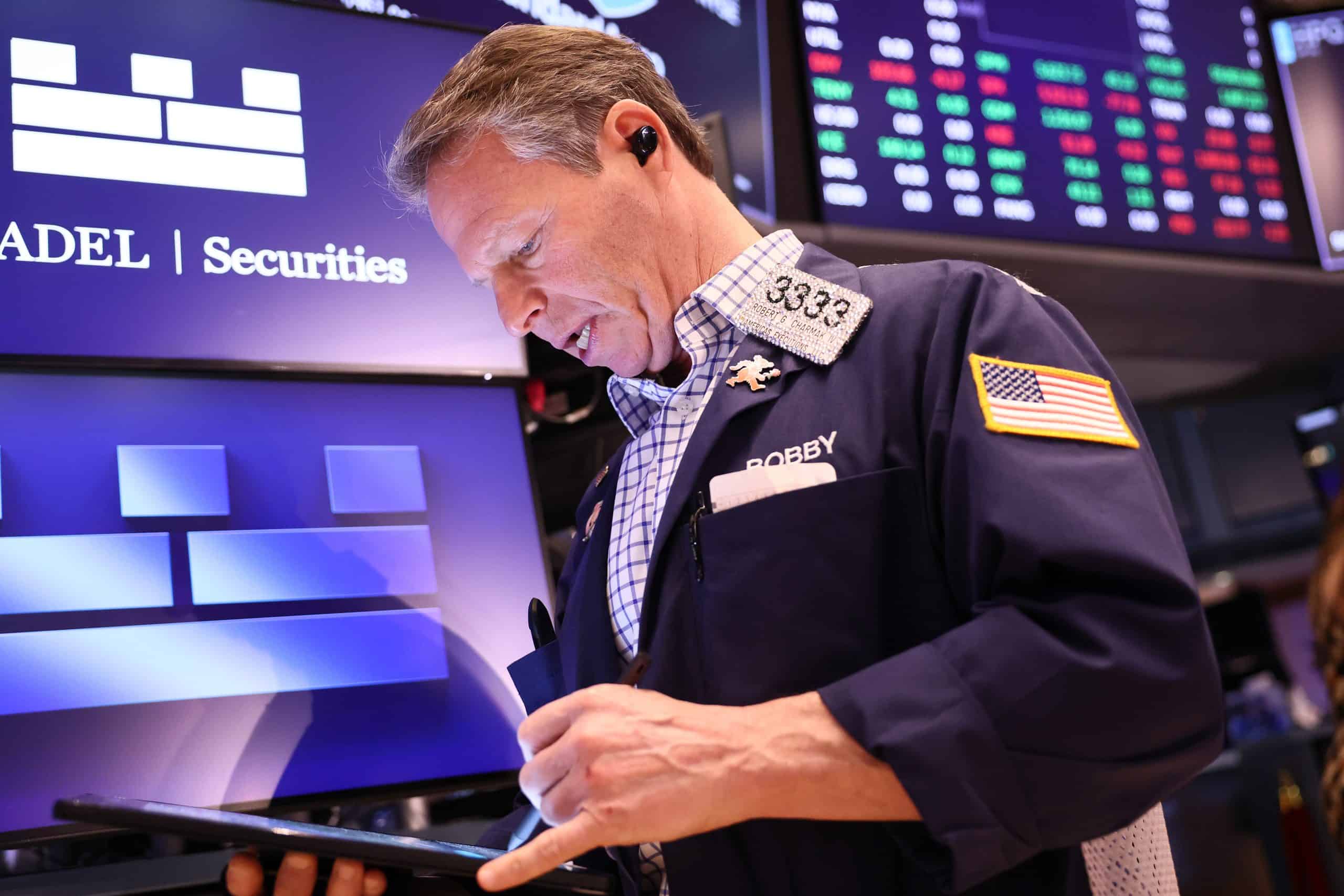 A male stock trader on the New York Stock Exchange floor looks down at a handheld device. He wears a dark blue jacket over a white and blue plaid shirt, an earbud in his right ear, and a name badge on his lapel reading '3333 Robert G. Charmax IV' and 'Bobby'. An American flag patch is visible on his left sleeve. In the background, blurred digital screens display market data with green and red figures and the 'ADEL | Securities' logo.
