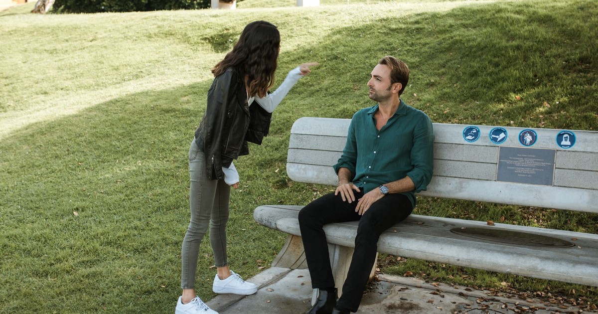 A couple in a heated argument while sitting and standing in a sunny park setting.