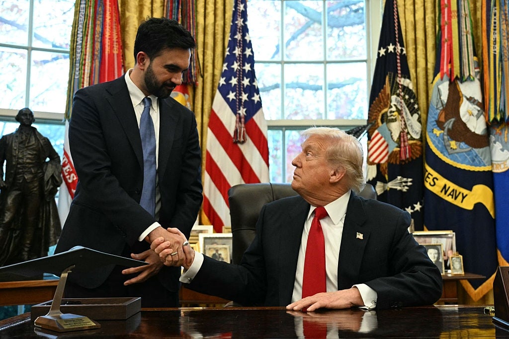 Two men in formal suits shake hands in an office with flags and an ornate background