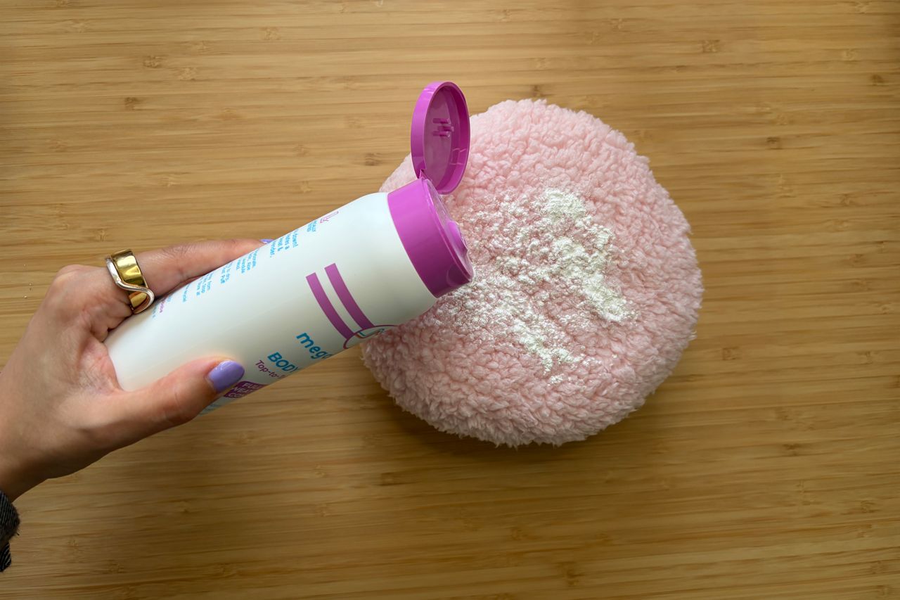 Hand pouring body powder from a container onto a round pink fluffy object wooden surface in the background