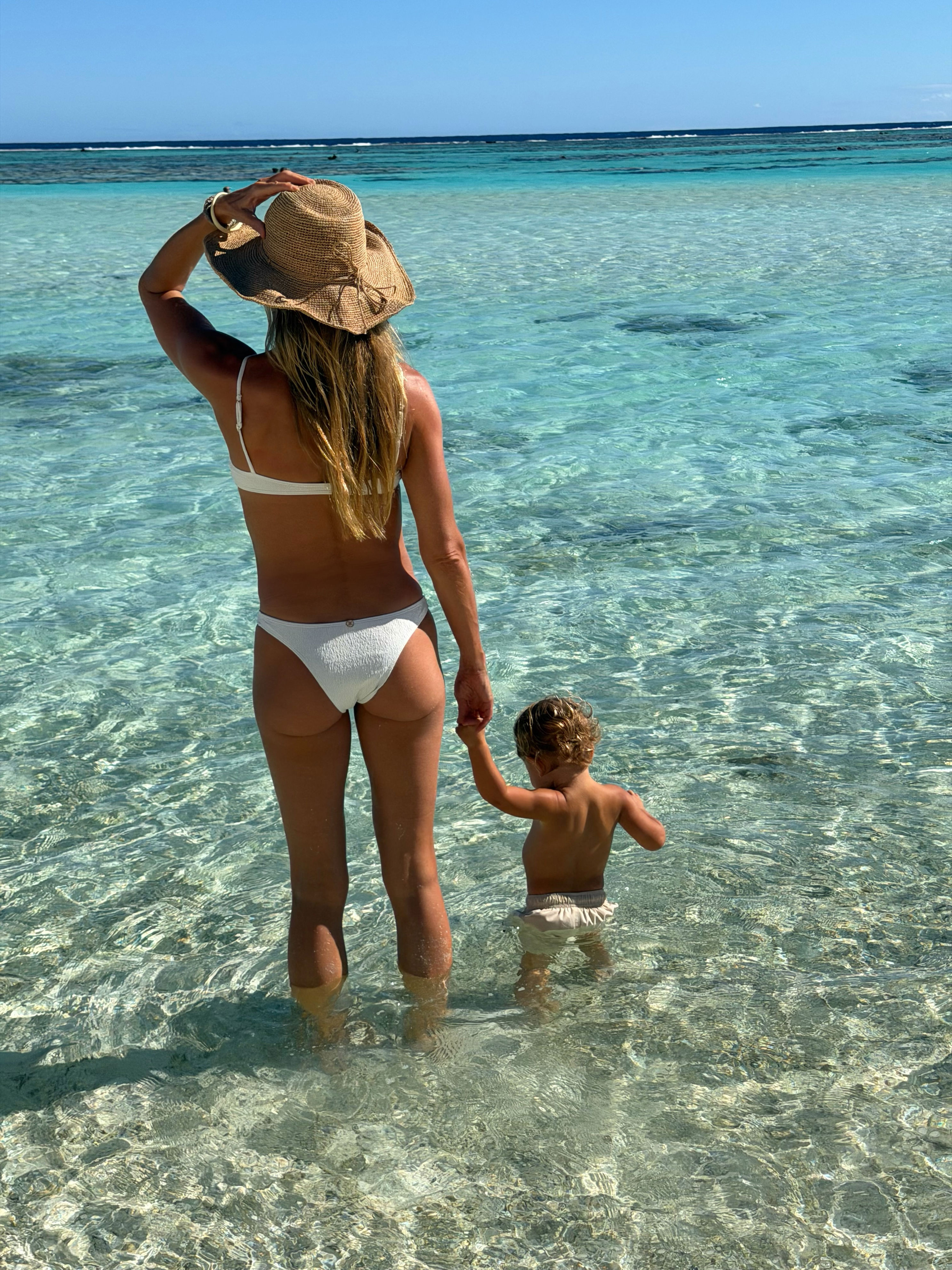 Gisele Bundchen stands in clear ocean water holding hands with a small child.