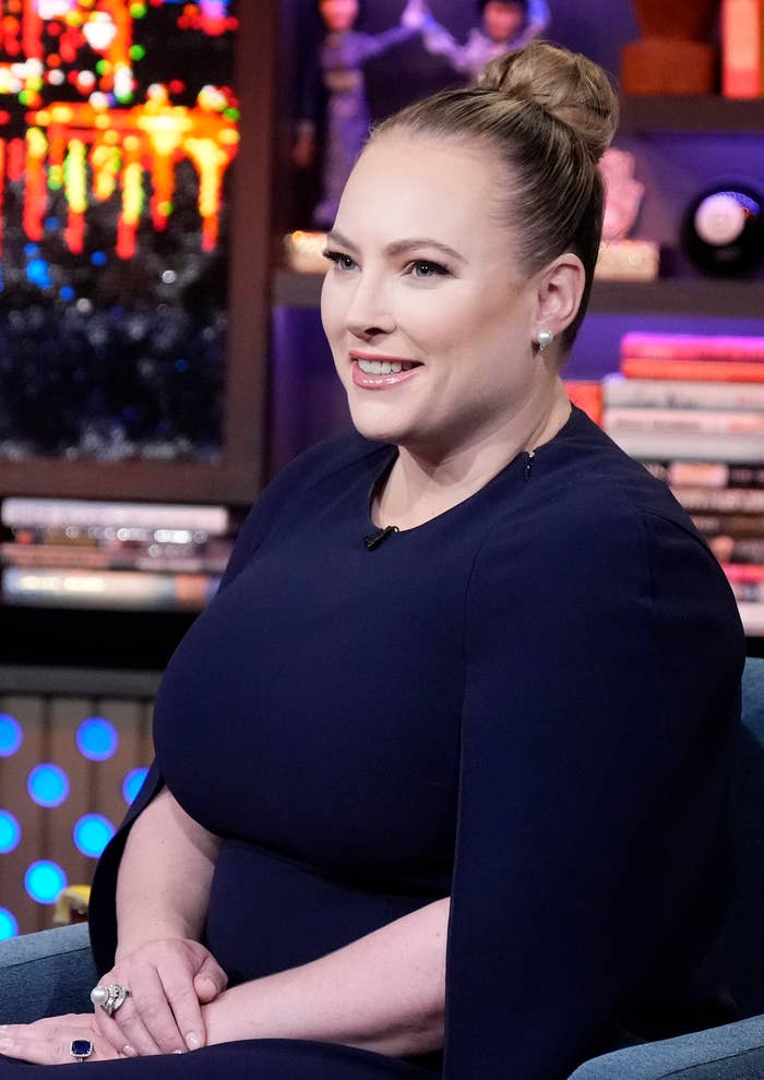 Person sitting on a talk show set, smiling, wearing a sleek, dark dress with a high bun hairstyle