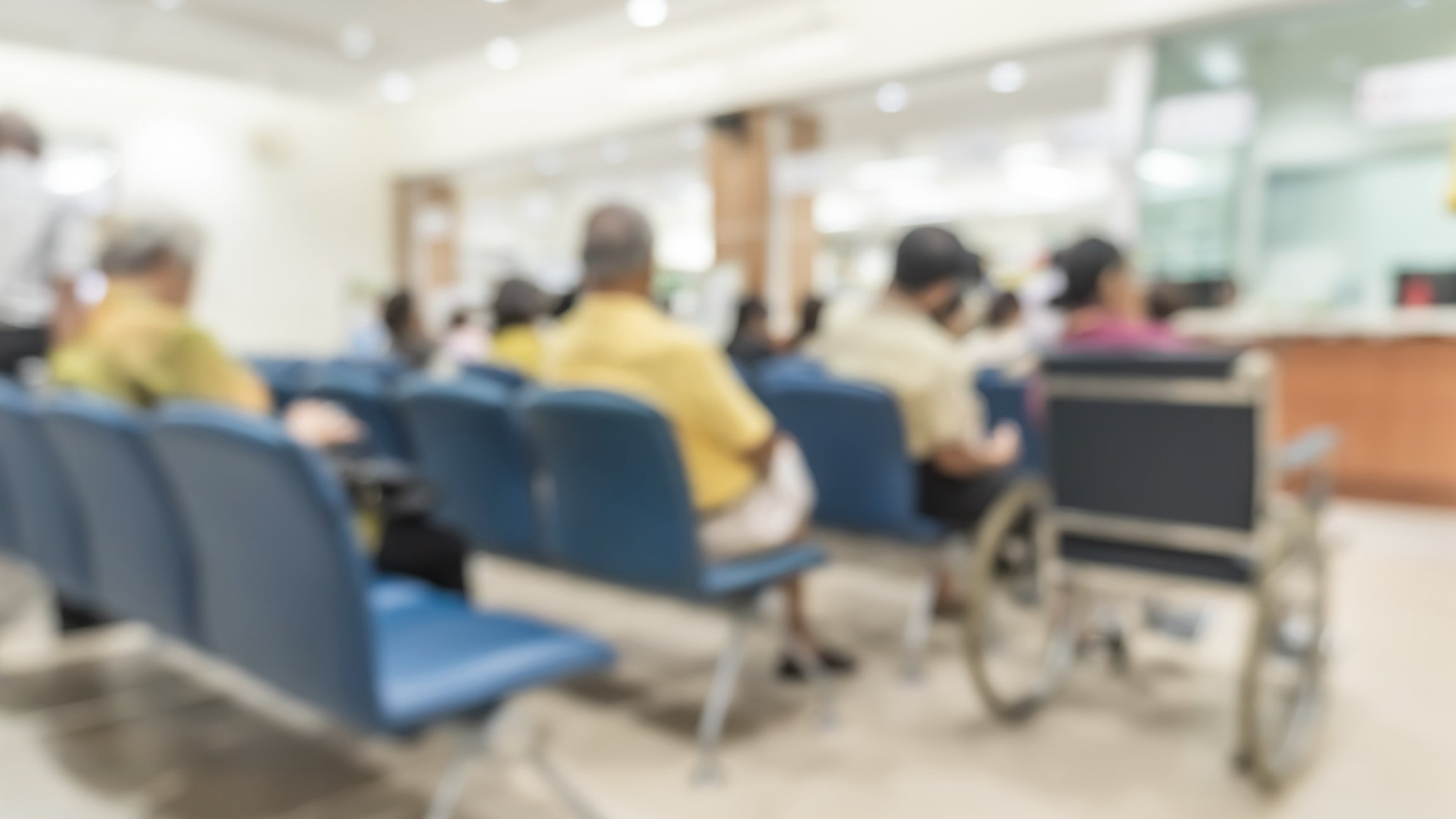 People seated in a waiting area, some in chairs, one in a wheelchair, facing a service counter in a public or medical facility