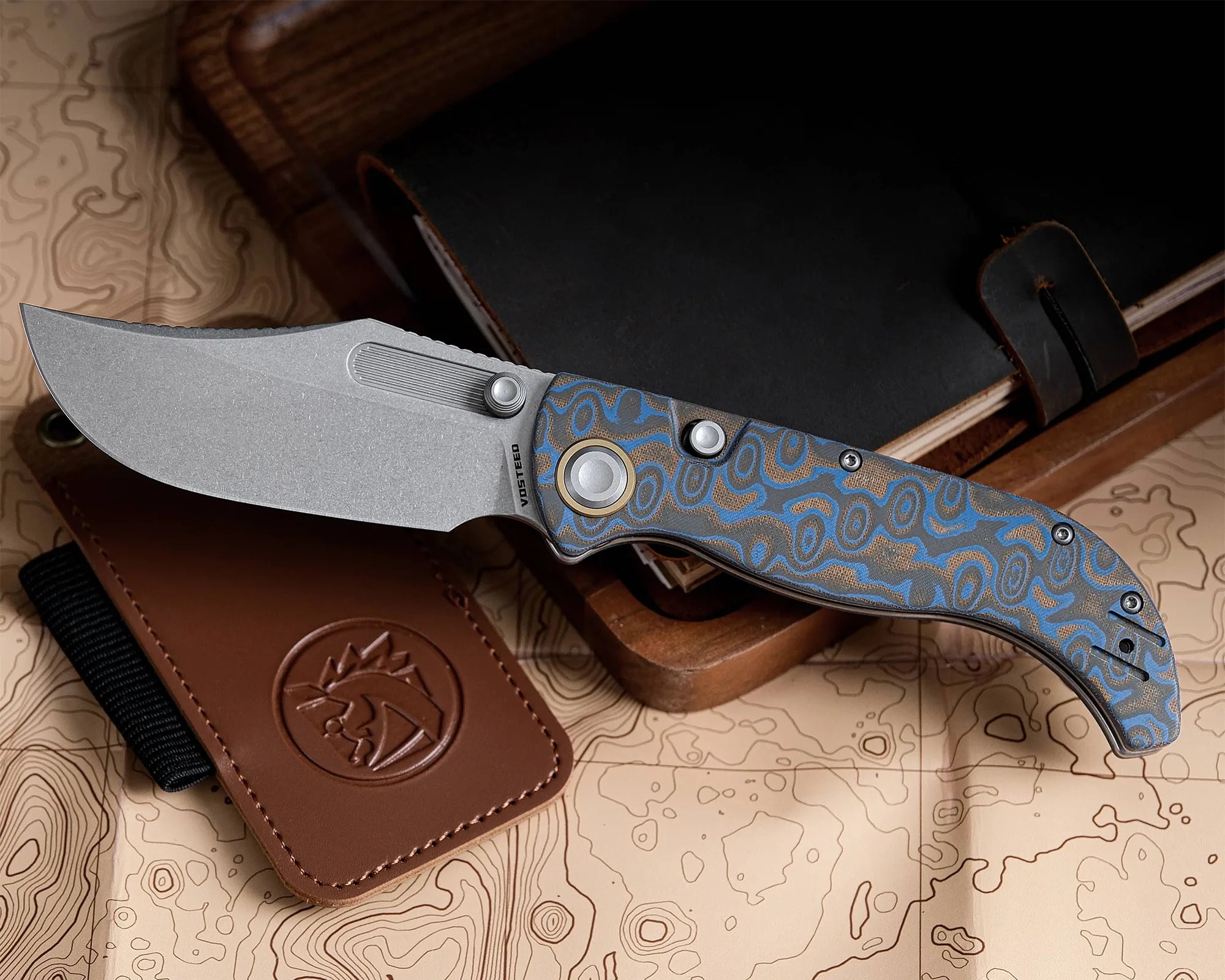 Folding knife with a stonewashed blade and blue patterned handle resting on a topographic map and leather accessories.
