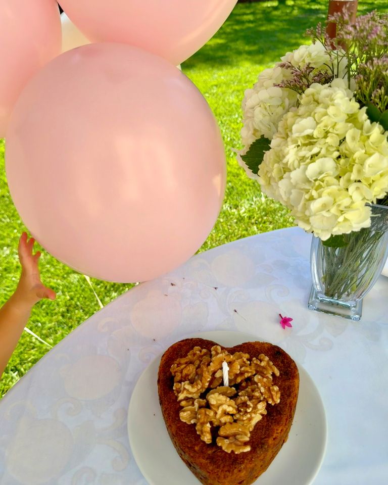 A heartshaped cake with walnuts and a candle on a plate pink balloons and flowers in the background childs hand reaching for a balloon