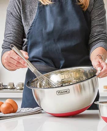 Person whisking in a metallic Viking mixing bowl on a kitchen counter with eggs nearby