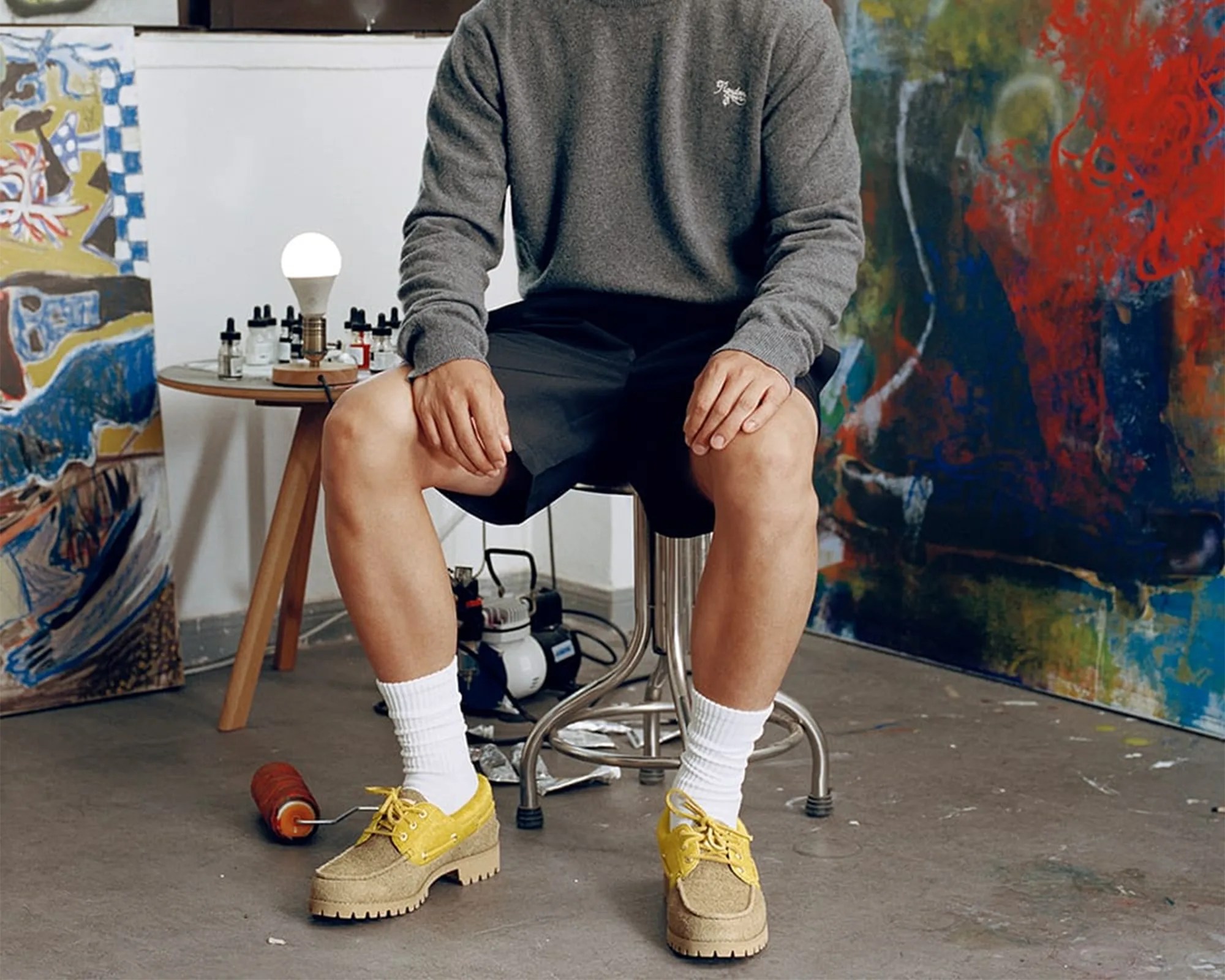 Person wearing yellow lace-up platform shoes with white socks, black shorts, and a gray sweater sitting on a metal stool in an art studio.