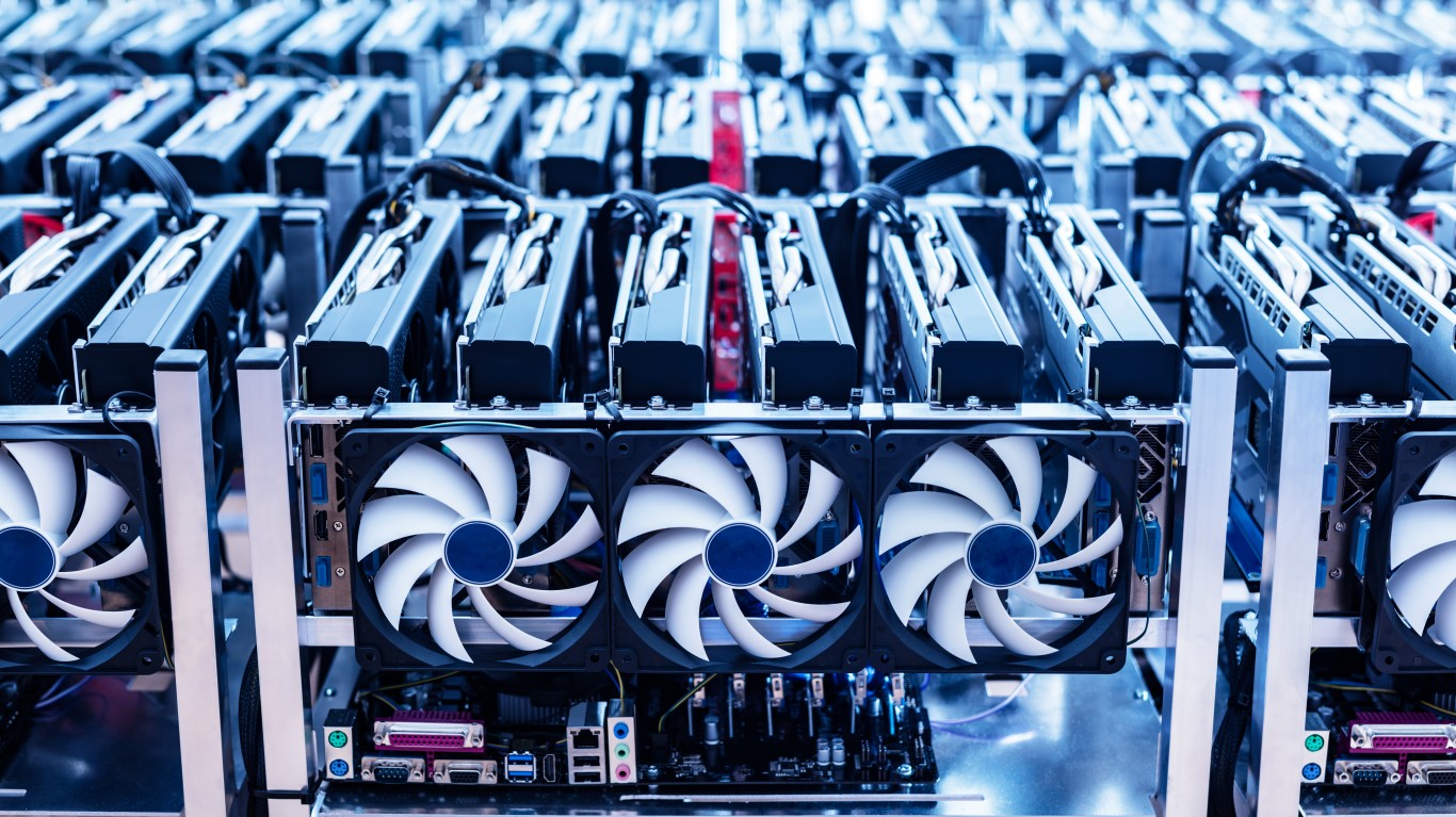 A close-up shot displays multiple rows of black graphics processing units (GPUs) with prominent white cooling fans, each featuring a dark blue center. The fans are arranged in neat, parallel rows, with various connecting cables visible between the units, set against a blurred background of more identical hardware in cool blue tones. The image conveys the dense and repetitive industrial nature of high-performance computing equipment.