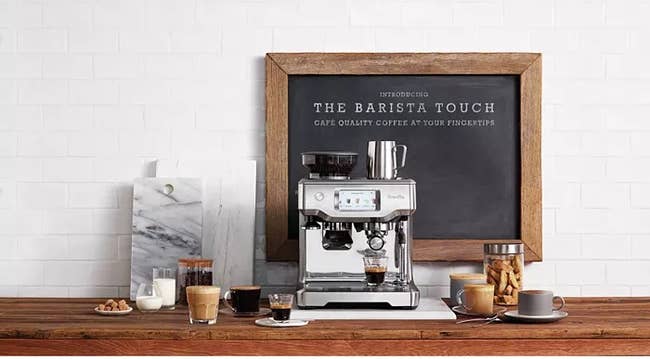 A sleek espresso machine on a kitchen counter, surrounded by coffee drinks and a framed chalkboard sign promoting