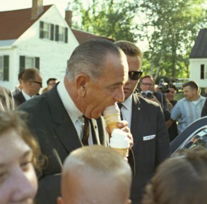 A man in a suit enjoys an ice cream cone among a crowd at an outdoor event
