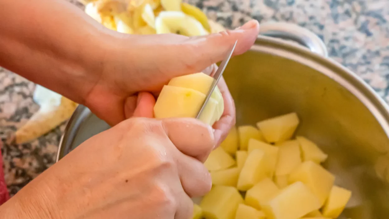 Woman hands cutting potatoes into chunks to prepare food in a metal pot.