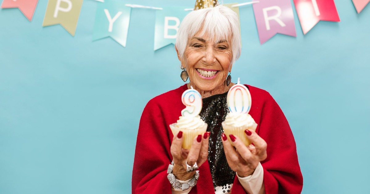 Smiling elderly woman holding cupcakes with candles on 90th birthday party.