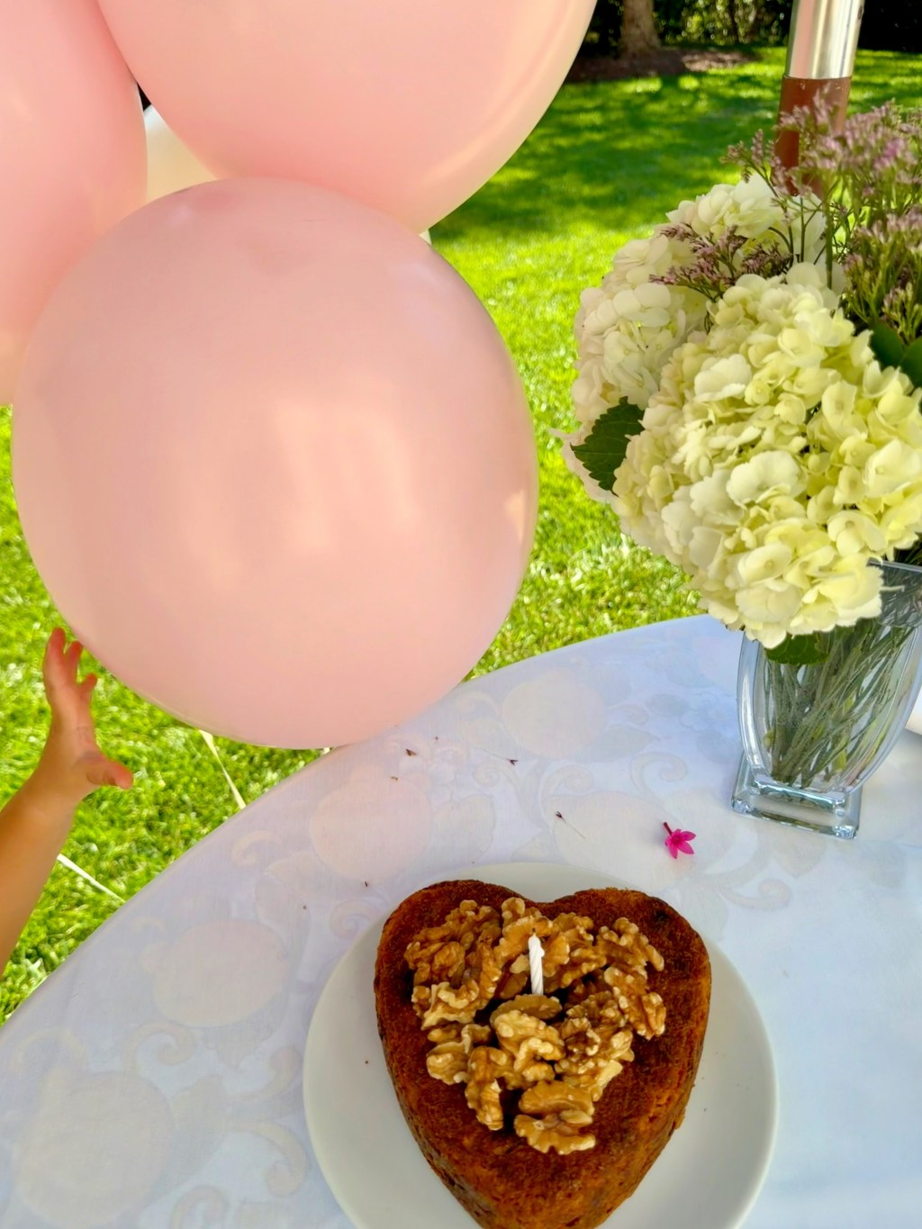 Heart-shaped birthday cake with walnuts, a candle, pink balloons, and white flowers on a white tablecloth, with a child's hand reaching for a balloon.