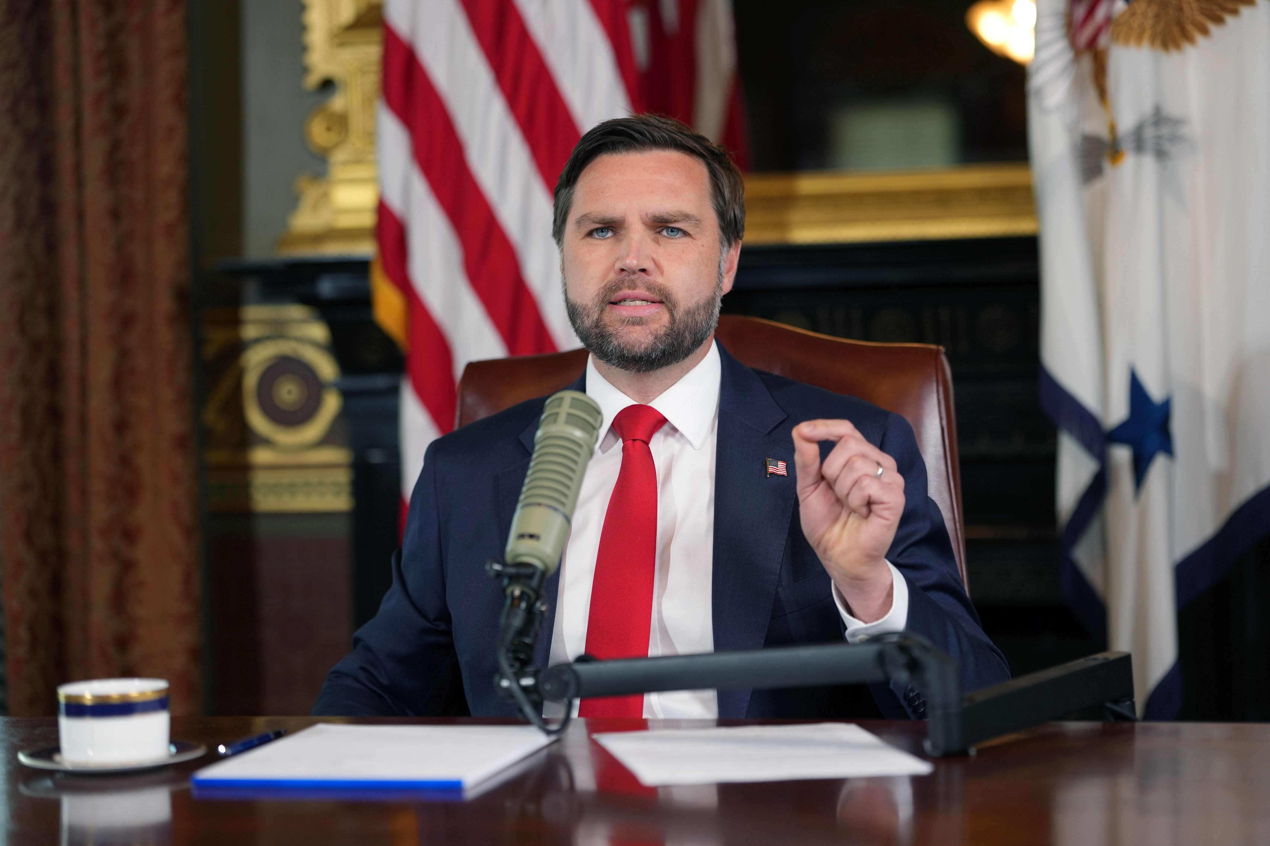 A person in a suit speaks at a desk with a microphone, in front of flags and a formal room setting, suggesting a press conference or official statement