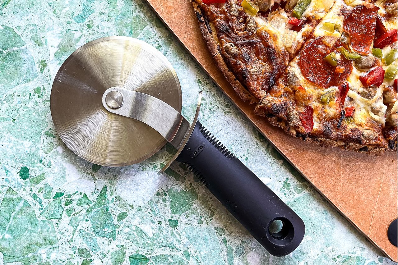 An OXO pizza cutter next to a slice of pizza on a cutting board