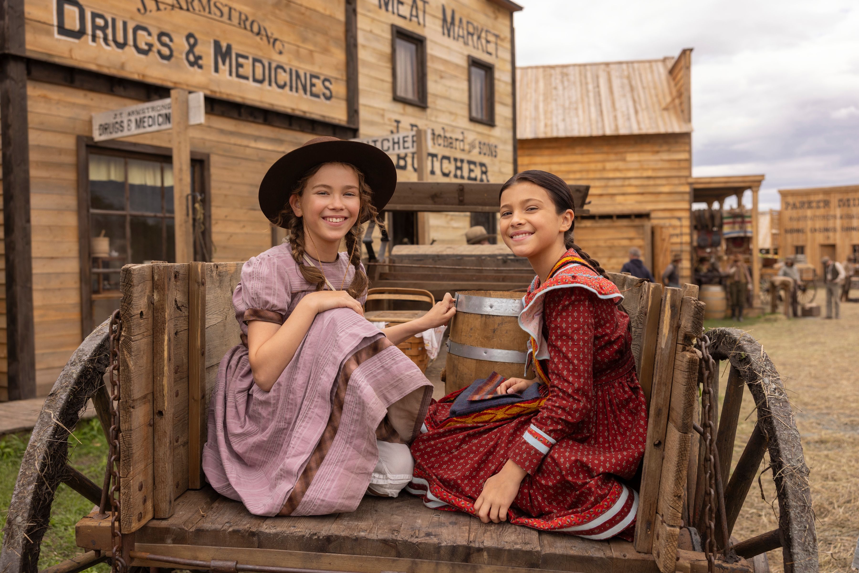 Alice Halsey as Laura Ingalls and Wren Zhawenim Gotts as Good Eagle sit in the back of a cart in town