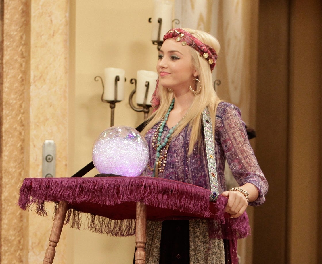 Two people in a living room: one in a white halter dress, the other in a fortune teller outfit with a crystal ball between them