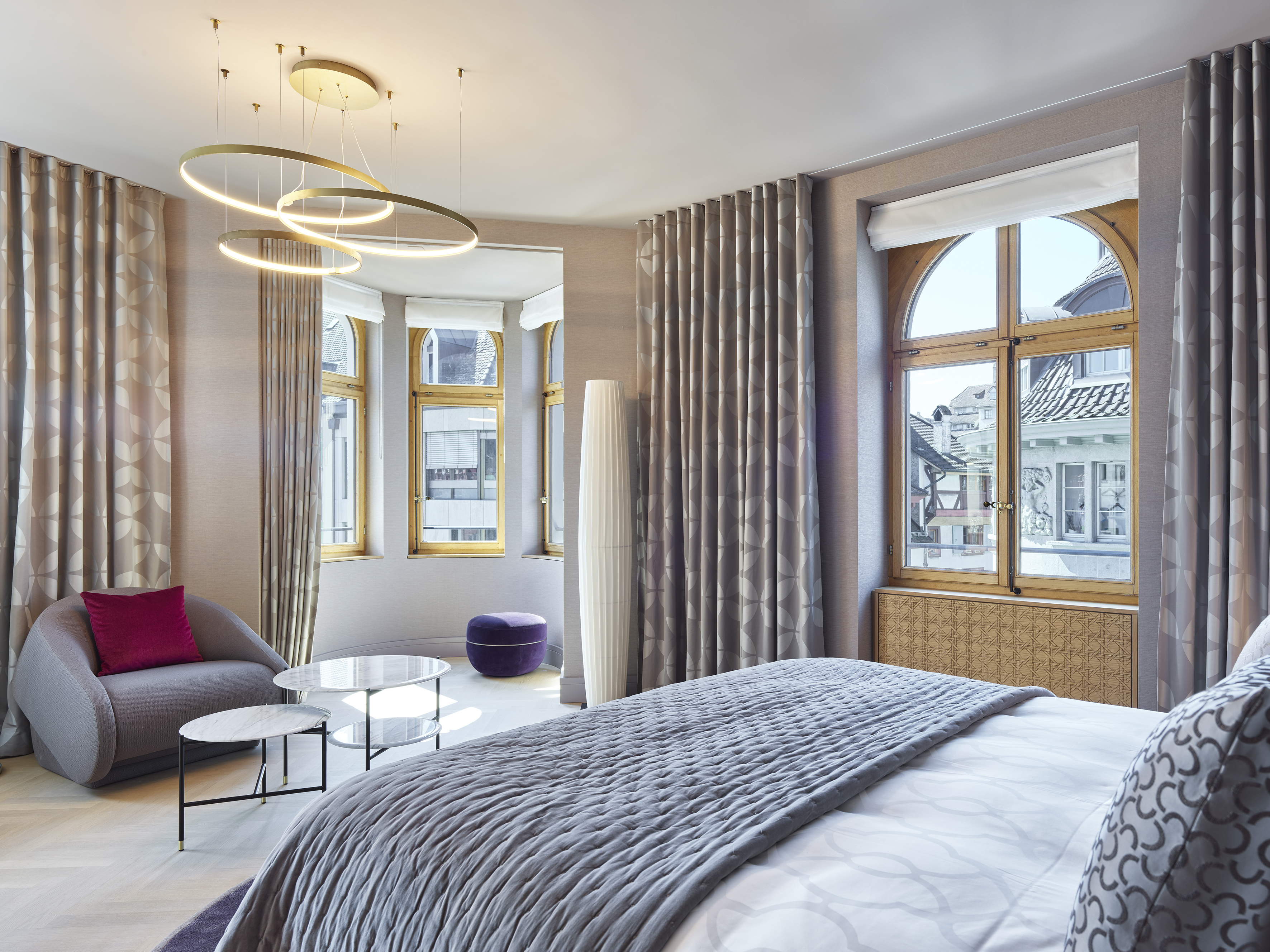 Photos of the interior of Hotel Märthof Basel shows a gray blanket covering a white bedspread, with retro style furniture and large arched windows.